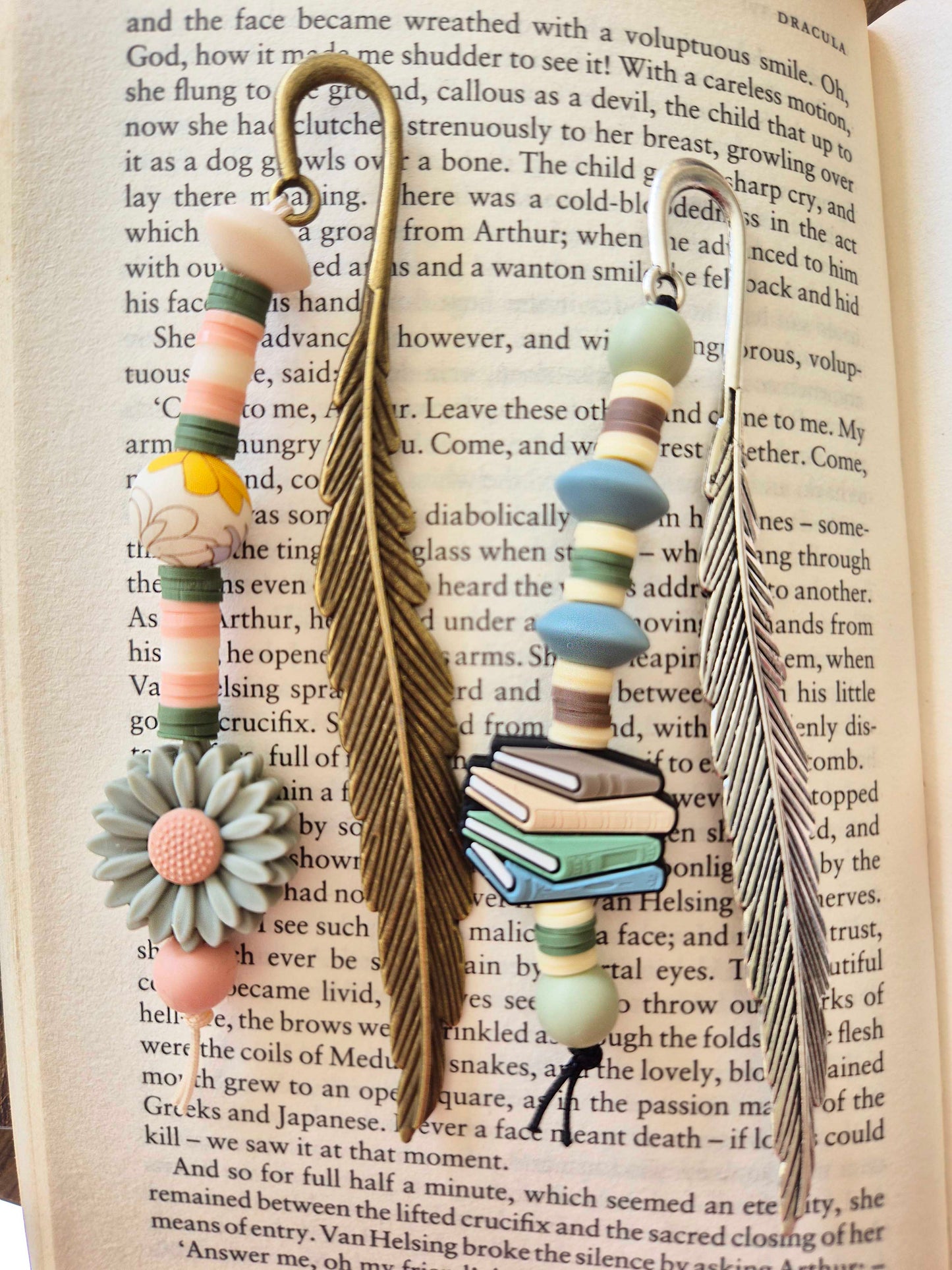Metal feather bookmarks decorated with beads. Bronze feather bookmark with green daisy focal bead. Silver feather bookmark with stack of books as a focal bead.