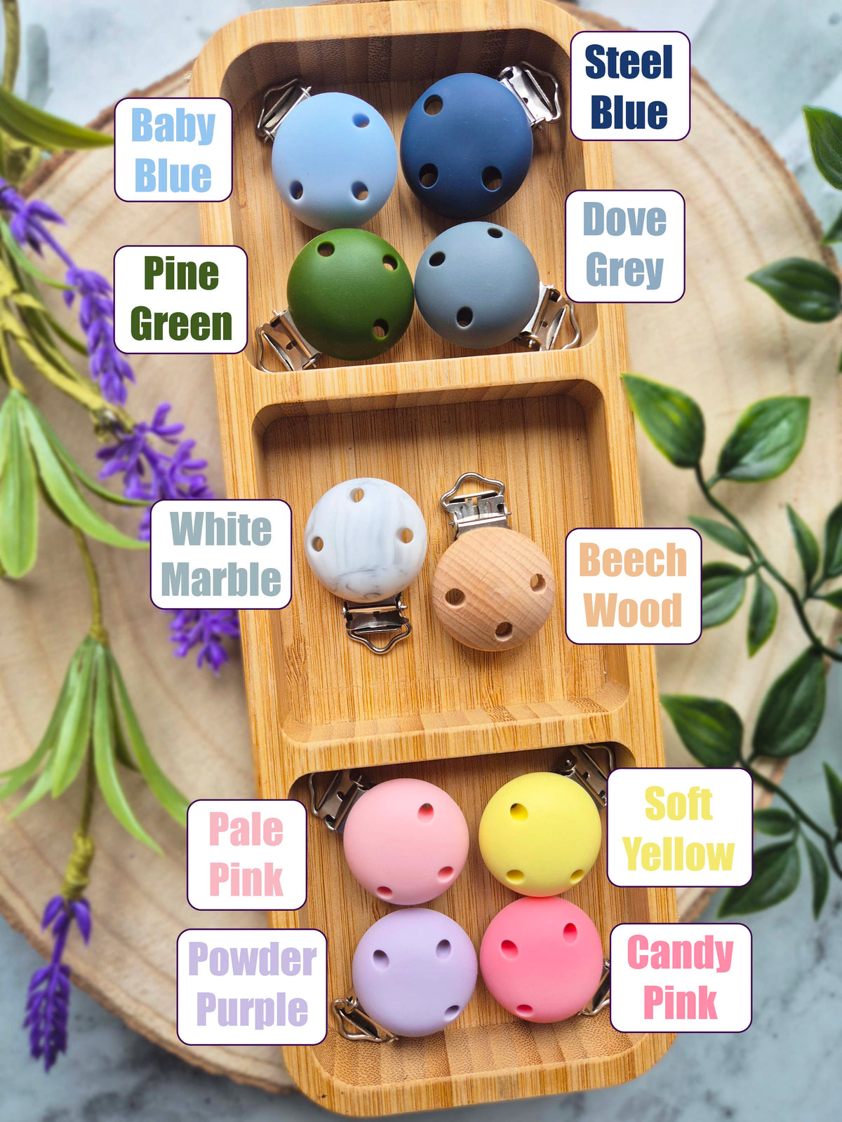 A wooden divided tray displays multiple wooden baby teething toys in different colors, with text labels identifying each color variant such as Baby Blue, Pine Green, White Marble, and Soft Yellow. The toys are arranged on a round wooden slice surrounded by decorative greenery.