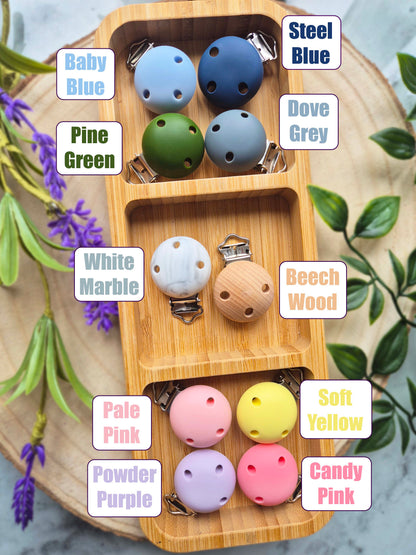 A wooden divided tray displays multiple wooden baby teething toys in different colors, with text labels identifying each color variant such as Baby Blue, Pine Green, White Marble, and Soft Yellow. The toys are arranged on a round wooden slice surrounded by decorative greenery.