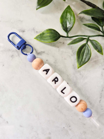 A personalized beaded keychain with a blue carabiner, blue and wood beads, and white letter beads spelling 'ARLO', displayed on a light marble surface with green leaves in the background.