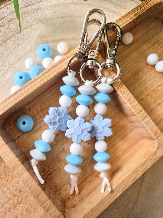 Three beaded keychains with silver lobster clasps, featuring light blue and white beads, snowflake charms, and white cords, displayed on a wooden tray.