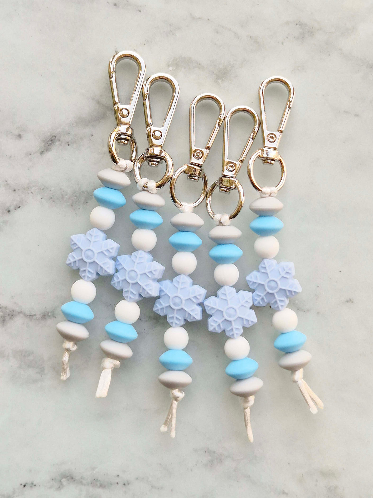 Four snowflake-themed keychain tags with light blue and white beads, silver-tone metal lobster clasps, and white cords, arranged on a light gray marble surface.