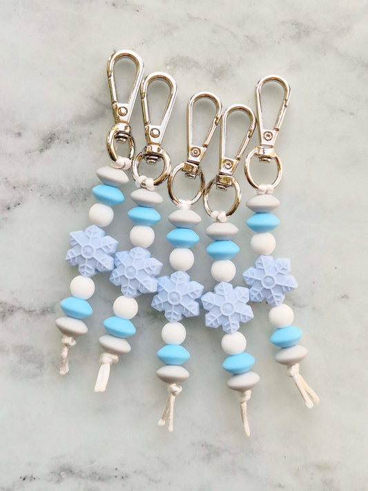 Four snowflake-themed keychain tags with light blue and white beads, silver-tone metal lobster clasps, and white cords, arranged on a light gray marble surface.