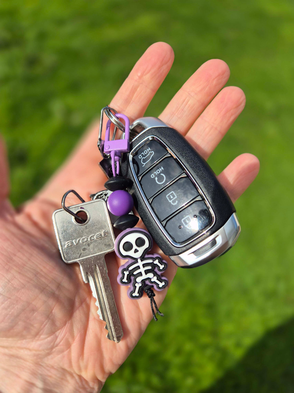 Skeleton keychain attached to a car fob and metal key. In a hand with grass blurred in the background