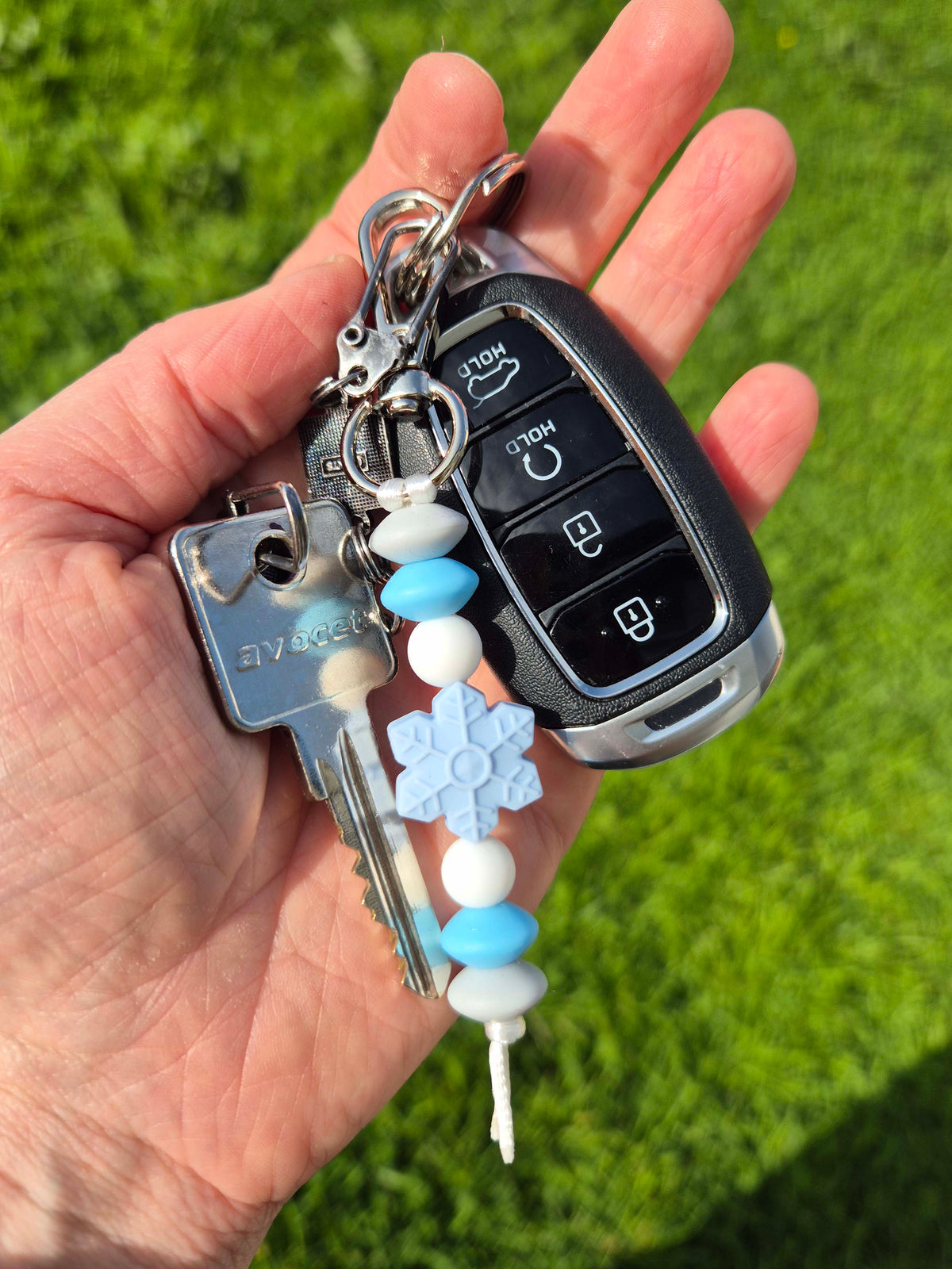 A hand holding a keychain with a car key fob, metal keys, and decorative blue and white beads, including a snowflake charm, against a blurred green grass background.