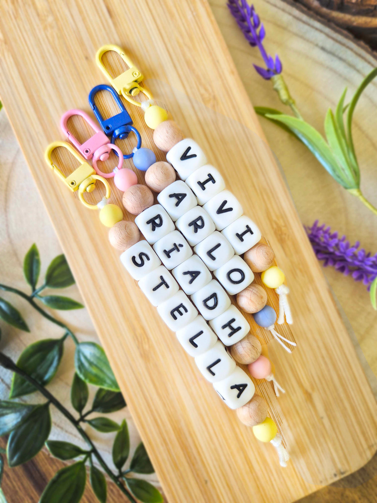 Three personalised beaded keychains with white letter beads spelling out names, displayed on a light wood board with decorative green leaves and purple flowers. Can be used as bag charms