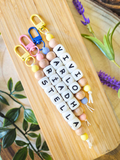 Three personalised beaded keychains with white letter beads spelling out names, displayed on a light wood board with decorative green leaves and purple flowers. Can be used as bag charms