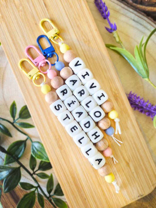 Three personalised beaded keychains with white letter beads spelling out names, displayed on a light wood board with decorative green leaves and purple flowers. Can be used as bag charms