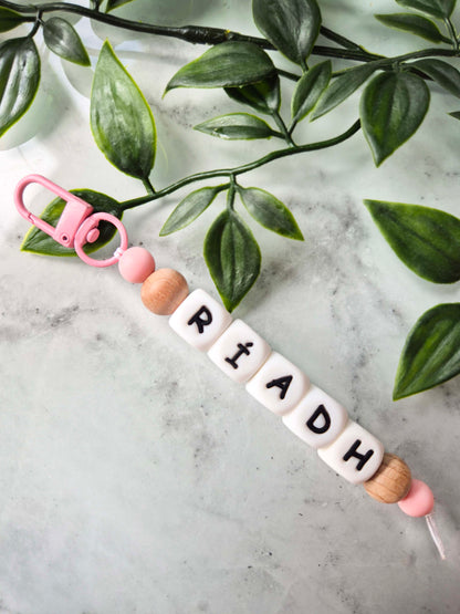 A personalised keychain or bag charm with a pink clasp, pink and wood beads, and white letter tiles spelling 'RIADH' on a light marble surface with green leaves.