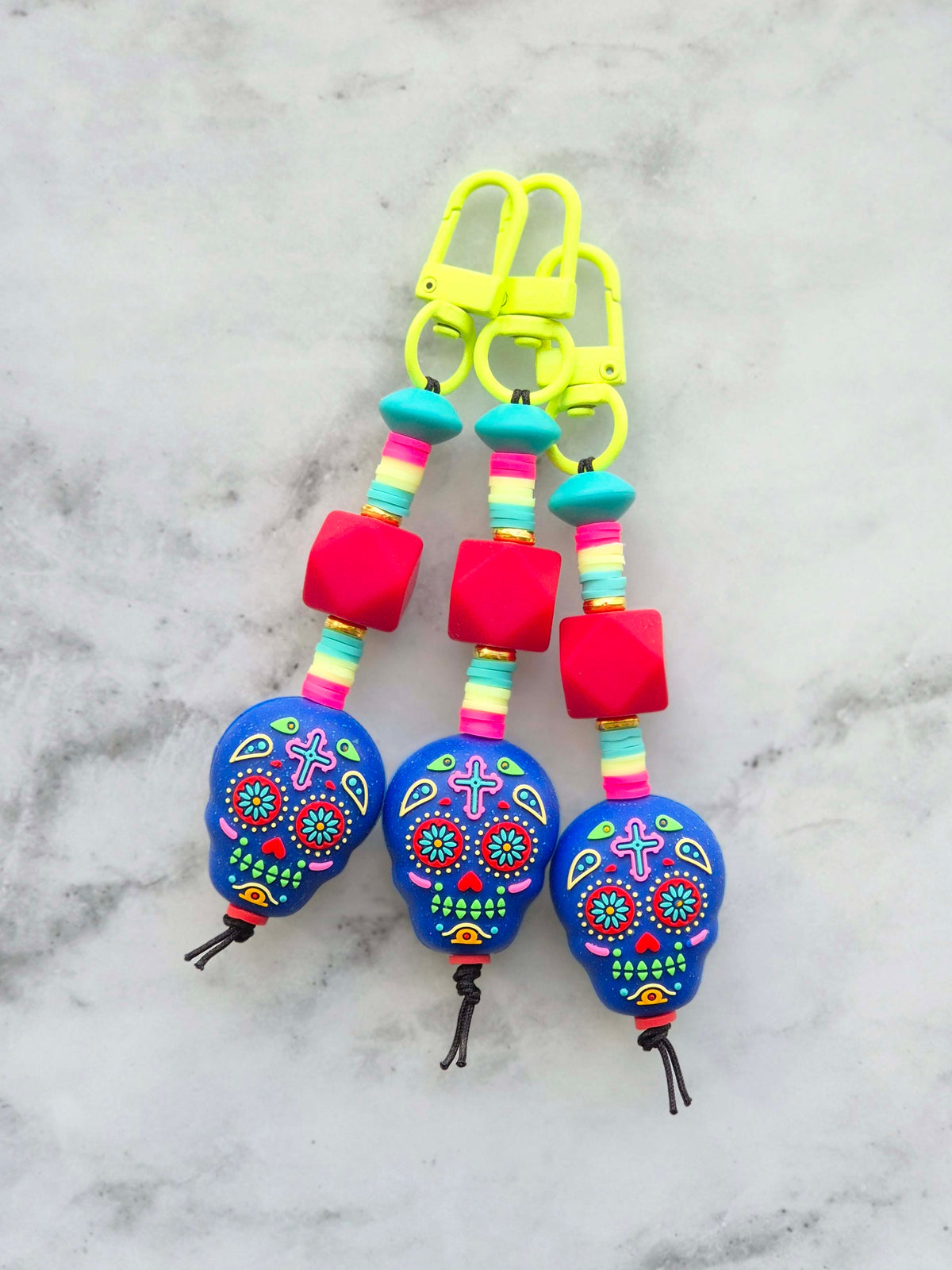 Three colorful sugar skull keychains with bright blue skulls, pink beads, and neon yellow clips, displayed on a light gray marble surface.
