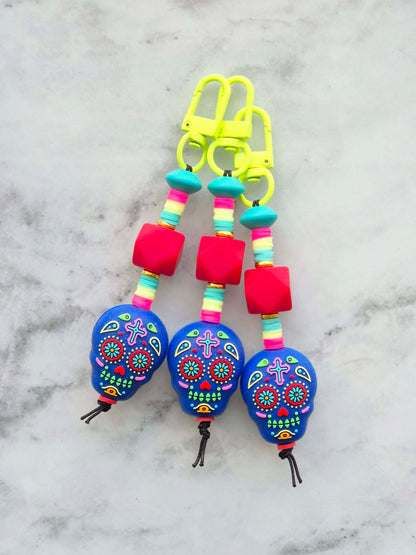 Three colorful sugar skull keychains with bright blue skulls, pink beads, and neon yellow clips, displayed on a light gray marble surface.