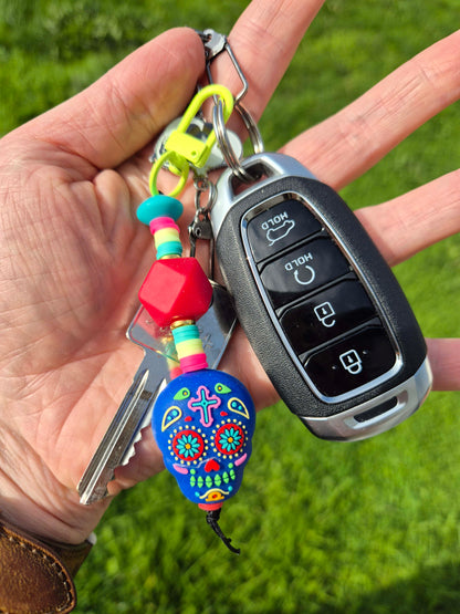 A hand holding a car key fob and keys outdoors on a green grass background. The keychain features a bright blue sugar skull charm with colorful floral details, a red and turquoise bead, and a neon yellow key ring.