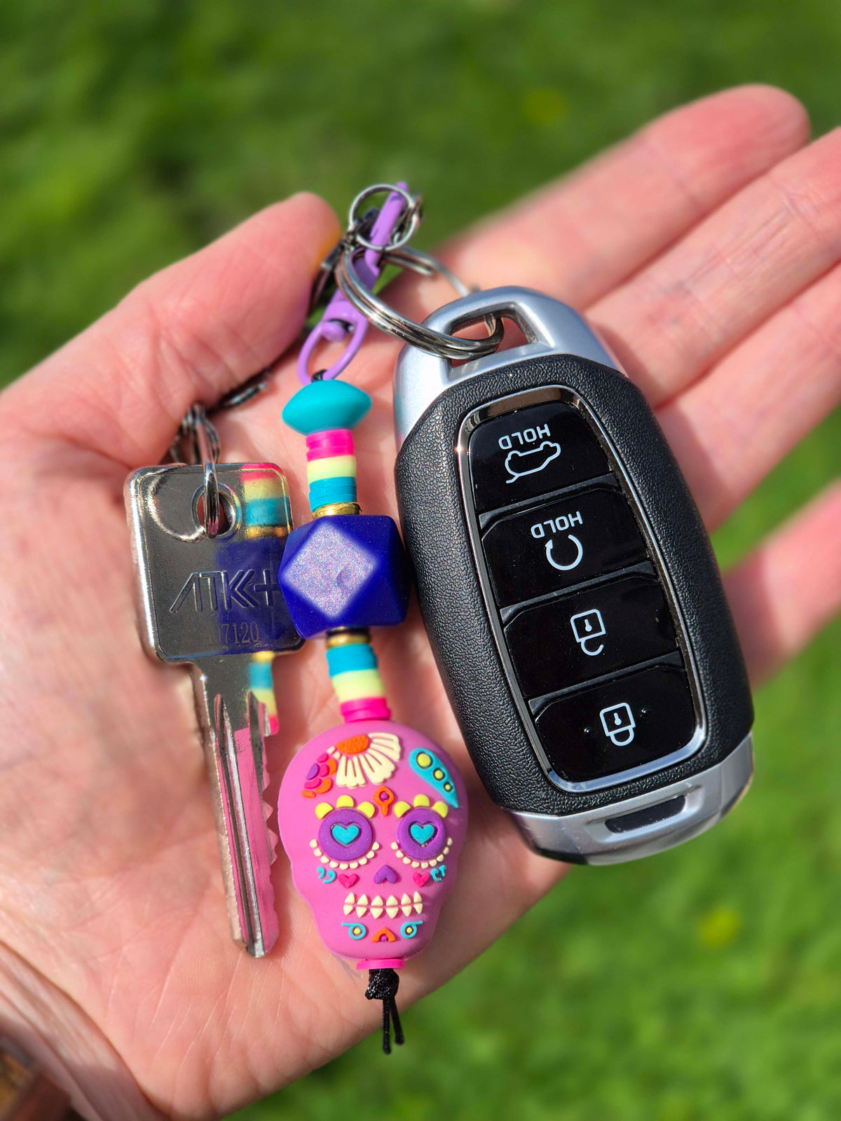 A hand holding a set of keys and a car key fob with colorful accessories, including a pink sugar skull charm, a blue hexagon keychain, and striped beads, against a blurred green grass background.