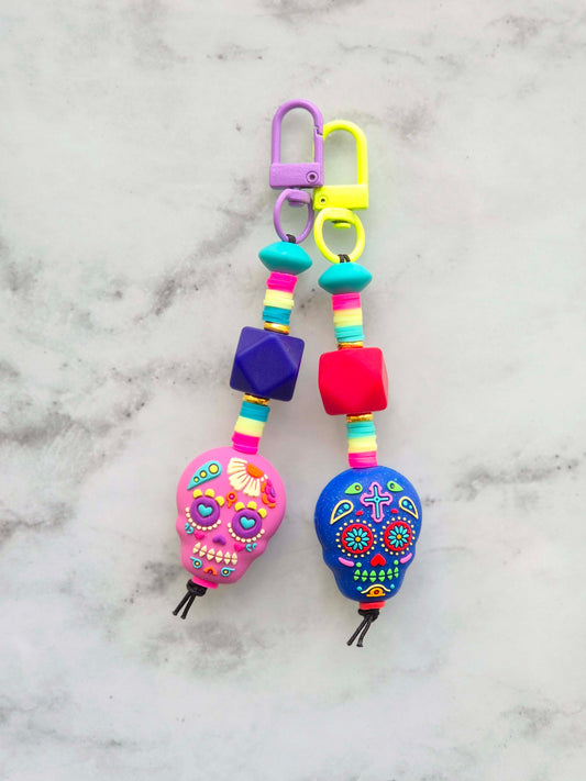 Two colorful beaded keychains with sugar skull charms, one pink and one blue, attached to purple and yellow carabiners, displayed on a light gray marble background.