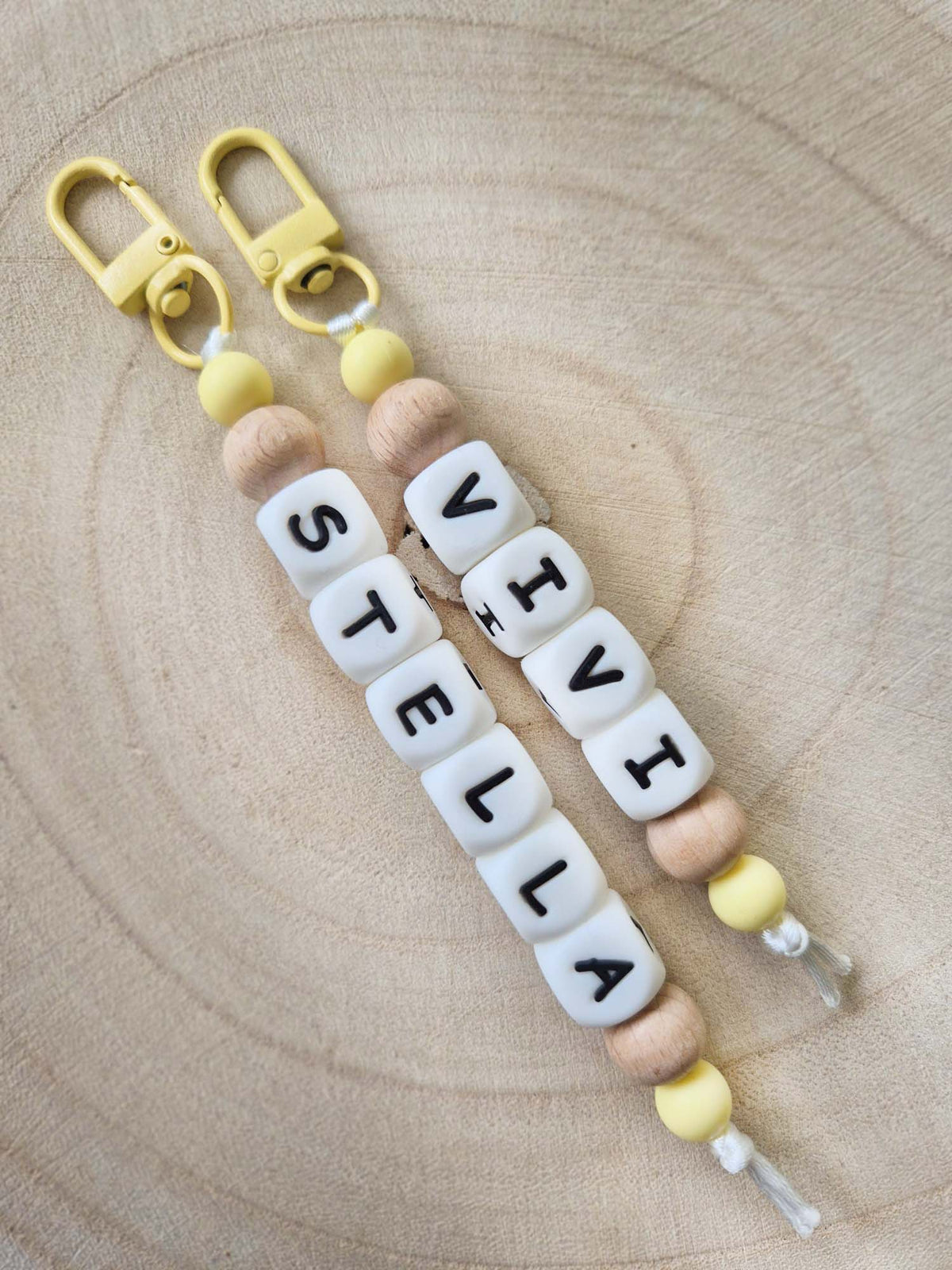 Personalised keychains / bag charms with the names Vivi and Stella written in while silicone beads. Made with yellow clips and yellow silicone beads and beech wood beads, displayed on a wooden board
